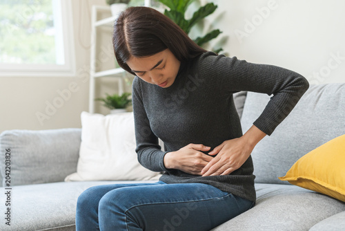 Valokuva Flatulence asian young woman hand in stomach ache, suffer from food poisoning, abdominal pain and colon problem, gastritis or diarrhoea sitting on sofa at home
