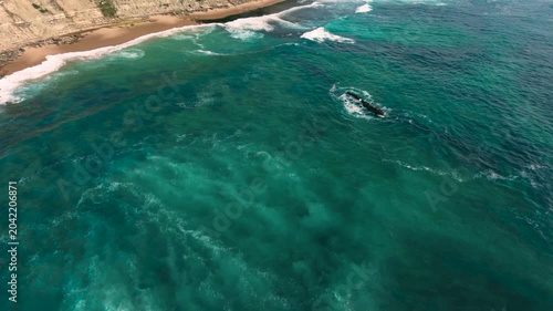 Aerial shot of the ocean with blue colours, waves doing  patters, rocks, sand and beach. Beautiful scenery