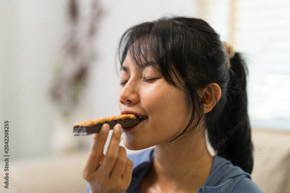 Ταπετσαρία Young Asian woman with a ponytail eats a healthy granola energy bar with her eyes closed, savoring a tasty nutritious wholesome snack in a bright comfortable home