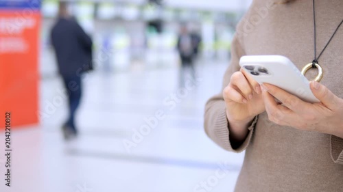 Close-up of woman holding phone in busy airport. She focuses on screen, calm and concentrated. Background shows travelers walking with luggage. Footage ideal for travel, communication