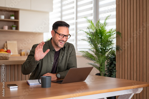 Adult man in smart casual clothes waving and smiling during a remote video call on his laptop from a bright home kitchen, staying connected for work or personal chats