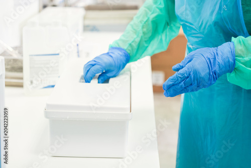 Medical worker in protective apron and blue gloves opens a sterile white plastic container at a clinic table, ensuring hygiene and safety standards in a professional lab environment