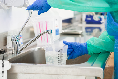 Gloved worker fills a graduated measuring pitcher with water in a clean medical facility, reflecting strict hygiene standards and professional preparation within a clinical environment