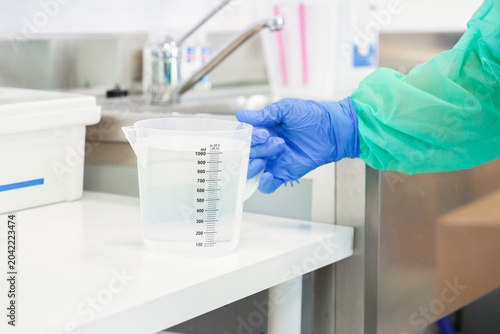 Gloved hand holding a clear plastic measuring jug with liquid in a professional laboratory, reflecting sterile scientific procedures and accurate chemical preparation in a research site