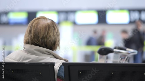 Woman waits in an airport terminal. She sits calmly, watching screens ahead. Her posture shows patience and anticipation. She wears a light jacket, scarf, and has short blonde hair