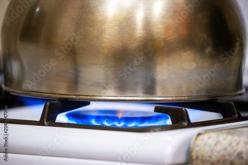 Metal kettle heating over a burning blue gas flame on a white kitchen stove. boiling water process, kitchen appliance usage