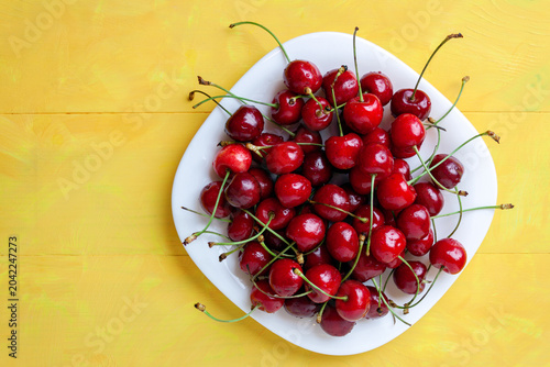 Fresh red cherries with green stems served on a white ceramic plate over a yellow wooden table. sweet summer berry,