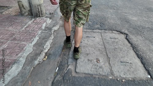Soldier in camouflage clothing walking along a dirty urban pavement and asphalt road. A first person perspective following the man's legs and feet as he moves forward on his patrol in the city
