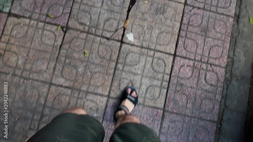 First person perspective of a man wearing sandals walking on a dirty urban sidewalk. The footage shows his feet and legs moving forward on patterned pavement, stepping past street litter