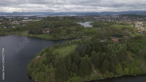  Aerial drone footage capturing the serene landscape of Stokka lake in Stavanger, Norway. The scene features a vast, calm body of water surrounded by lush greenery, captured at midday.