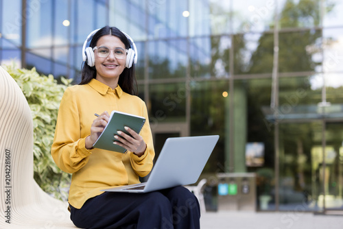 Foto Young female student smiling on a campus bench, wearing headphones, typing on a