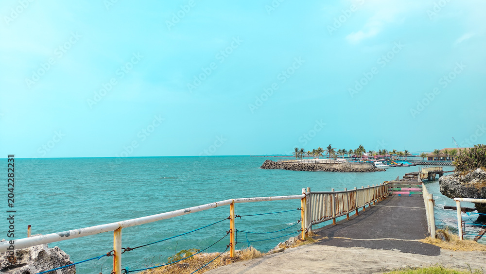 Fototapeta premium A rustic coastal walkway with white railings leading toward a tropical island with palm trees, surrounded by calm turquoise ocean waters under a clear sky in a peaceful summer setting