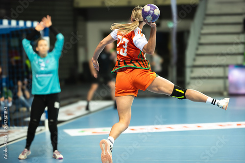 Fotografie A female handball player in an orange jersey jumps to throw the ball towards the goal defended by a goalkeeper in blue