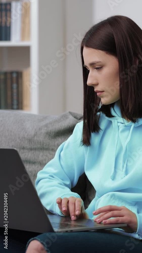 Woman sits on couch using laptop, focused on work in a cozy living room setting.