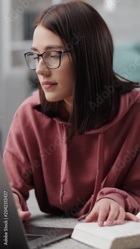 Woman studies at home using laptop while reading a book and taking notes.