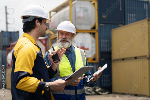 Foto Senior and young male workers in hard hats and safety vests review logistics data on a tablet at a busy container terminal, ideal for industrial and supply chain content