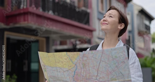 Portrait of woman traveler reads map and smiles standing in old downtown. Female with backpack observes landmarks as holds city plan against ancient buildings