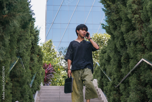 Canvas Print Confident young asian man talking on cell phone while walking down modern outdoor stairs next to green hedge and building