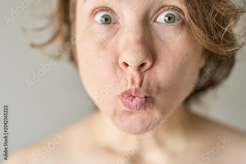 фотографія Woman Puckering Lips With Wide Open Eyes Making Playful Surprised Face In Neutral Studio Setting Closeup Portrait Bare Shoulders Under Bright Natural Light
