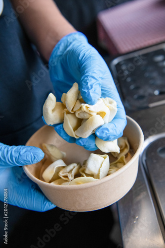 Food service worker wearing blue gloves handling uncooked stuffed pasta in a takeaway bowl, highlighting hygiene, food preparation, freshness, and commercial kitchen standards.