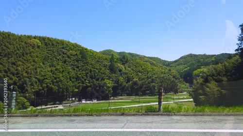 Scenic view of lush green rolling hills and traditional rural houses in Kumamoto prefecture during late spring. The point of view moves along a countryside road on bright and clear afternoon in April