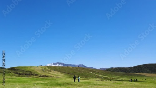 Wide shot of Kusasenri plateau at Mount Aso in Japan during spring season. People walk along dirt paths on fresh green hills under a clear blue sky in April. 