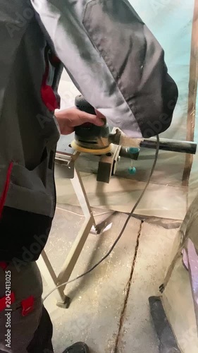 A worker in overalls is grinding a metal part using an orbital grinder on a workbench.