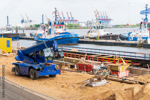 Schilderij op canvas Blue telescopic handler stands on a sandy construction site near harbor water, with tugboats, cranes, cargo terminals, building materials, formwork, and work equipment visible around the area