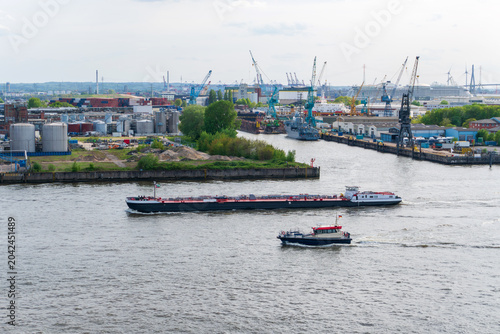 Photography Long cargo barge and small service boat travel through a wide harbor channel with shipyards, cranes, tank farms, warehouses, and industrial docks in the background