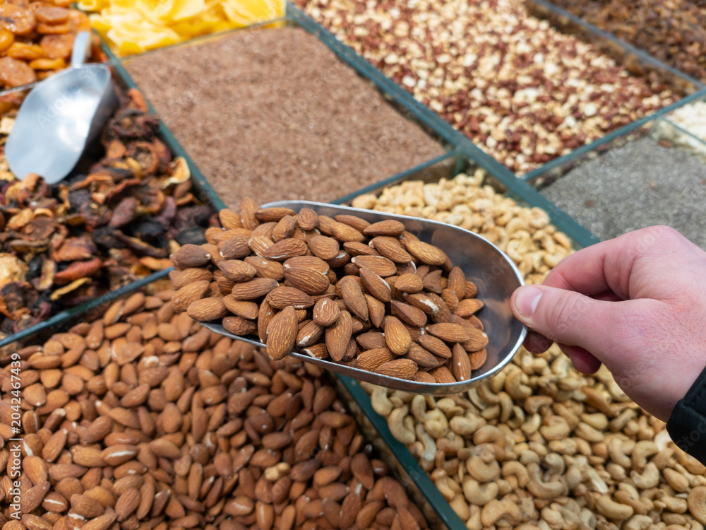 Obraz premium A metal scoop with almonds in a customer's hand over a store counter. The process of choosing fresh nuts at the market symbolizing quality food traditions and a healthy lifestyle