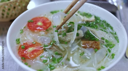 Close up of a person using chopsticks to eat a hot bowl of traditional vietnamese jellyfish noodle soup, a popular seafood dish with fresh vegetables, pork, and a savory clear broth