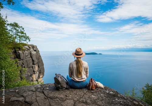 Back view of a woman in a hat sitting on a rocky cliff edge, looking out at a vast blue ocean with a small island and distant mountains under a cloudy sky