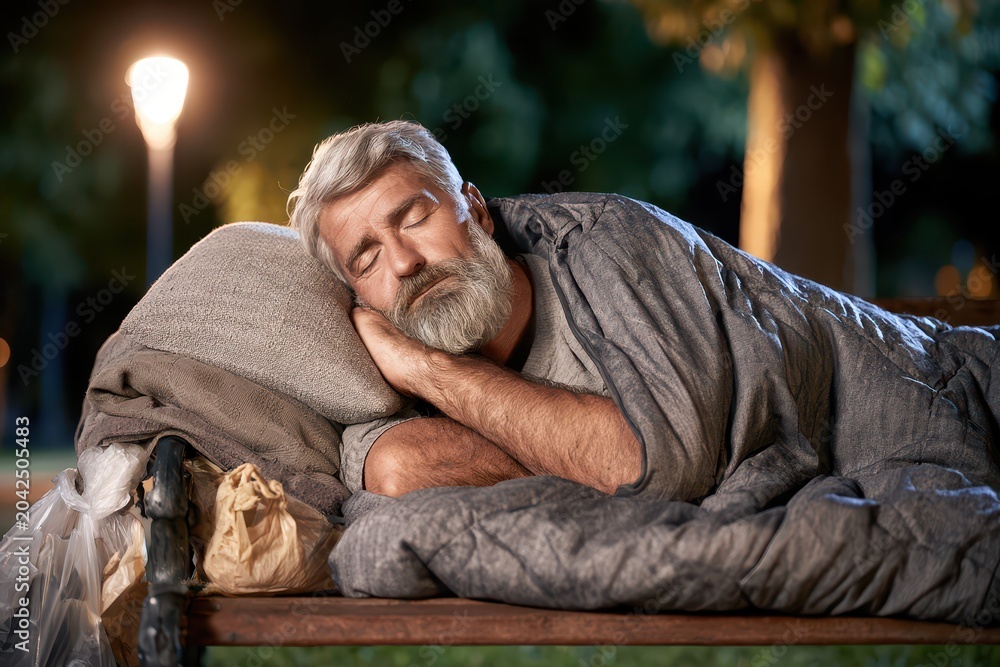 Fototapeta premium Elderly homeless man with grey beard sleeping on a park bench at night under a blanket.
