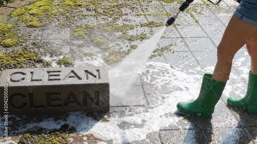 Person cleaning mossy patio pavers with a high pressure water sprayer, close up on boots and concrete block with the word clean engraved