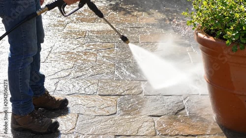Person using a pressure washer to clean a stone patio surface near a large terracotta plant pot