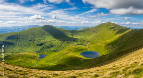 Majestic green mountain landscape featuring crystal clear alpine lakes under a bright blue sky with soft white fluffy clouds above