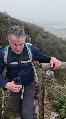Two male hikers climbing a steep, rocky trail on a foggy day. The older man stops at a viewpoint to take a selfie with his phone
