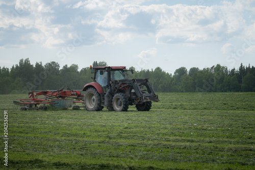 Bild auf Leinwand Tractor raking hay in green rural farm field under cloudy summer sky