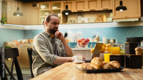 Konstfotografi Adult male frowning at laptop screen being disoriented while typing on laptop, uncertain man dealing with his remote work at kitchen table, Ambiguous pensive freelancer telecommuting