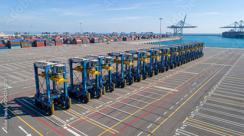 Symmetrical row of automated straddle carriers parked neatly inside vast empty cargo seaport shipping terminal