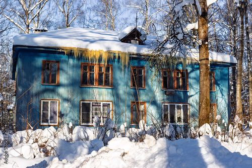 Charming weathered wooden house painted blue with heavy snow caps and long frozen icicles hanging from the roof during a bright and sunny winter day in the rural countryside