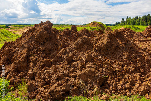 Large pile of raw dark soil mounds excavated from a field during construction work under a bright cloudy summer sky, showing natural clay and earth texture in an open landscape