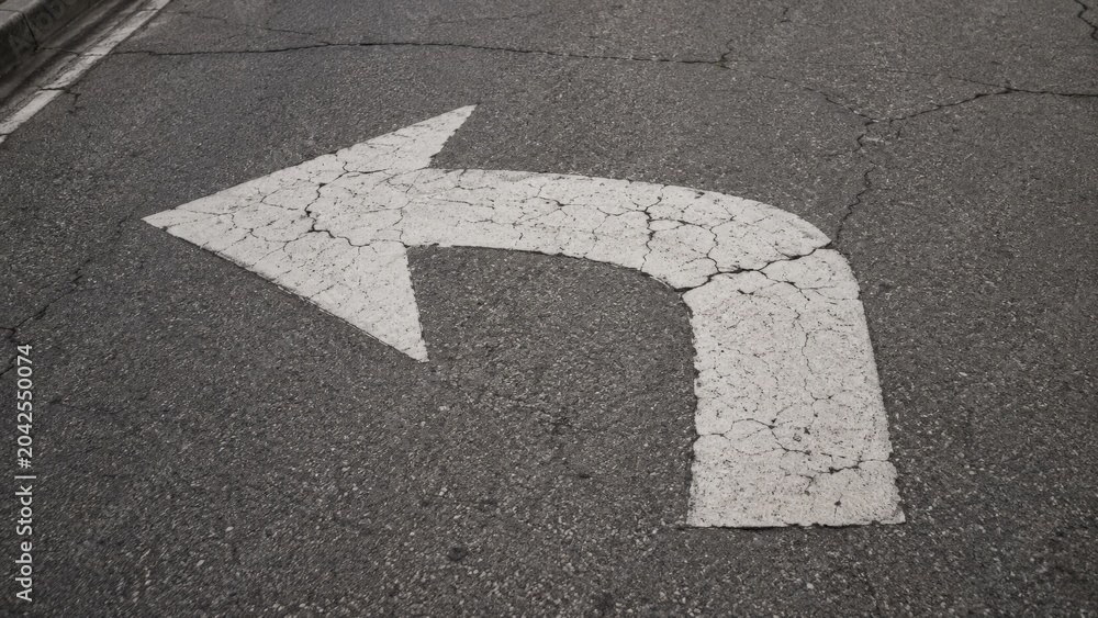 Fototapeta premium White painted left turn arrow on cracked asphalt road surface.