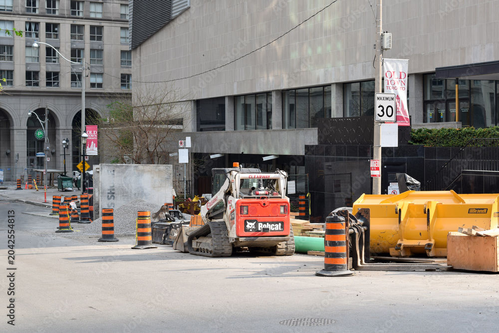 Naklejka premium Urban road construction site in downtown Toronto featuring a compact track loader, traffic barrels, construction materials, and street infrastructure improvement work, May 2026