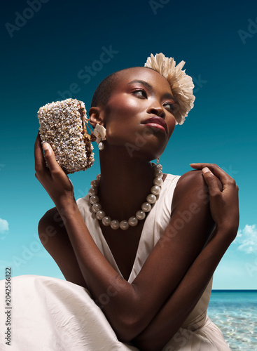 Editorial fashion portrait of gorgeous photo model wearing avant garde white haute couture dress with luxury pearl necklace and shell clutch on wooden pier at tropical turquoise summer beach