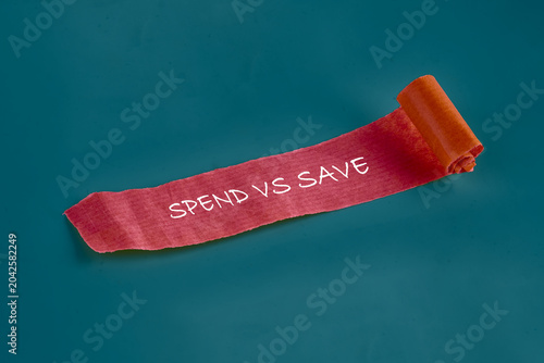 фотографія A vibrant roll of ribbon displays the phrase spend vs save on a teal background,