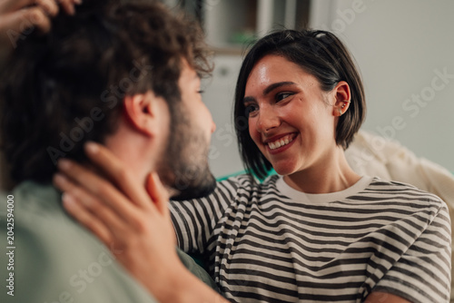 Young woman smiling at her partner while gently touching his neck, sharing a loving moment at home. Intimacy and connection