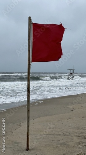 Red warning flag on a wooden pole stands on a windy beach, rough waves roll under a gray sky, distant lifeguard tower suggests hazardous surf