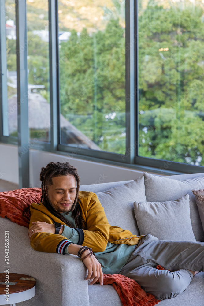Fototapeta premium African American man reclining on sofa in living room, wearing mustard jacket, showing smartwatch