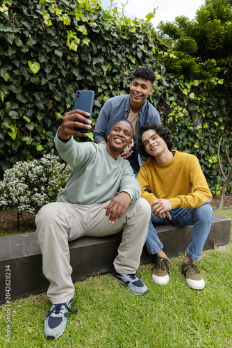Three teenage males sitting on low bench in backyard, wearing casual clothes and holding smartphone